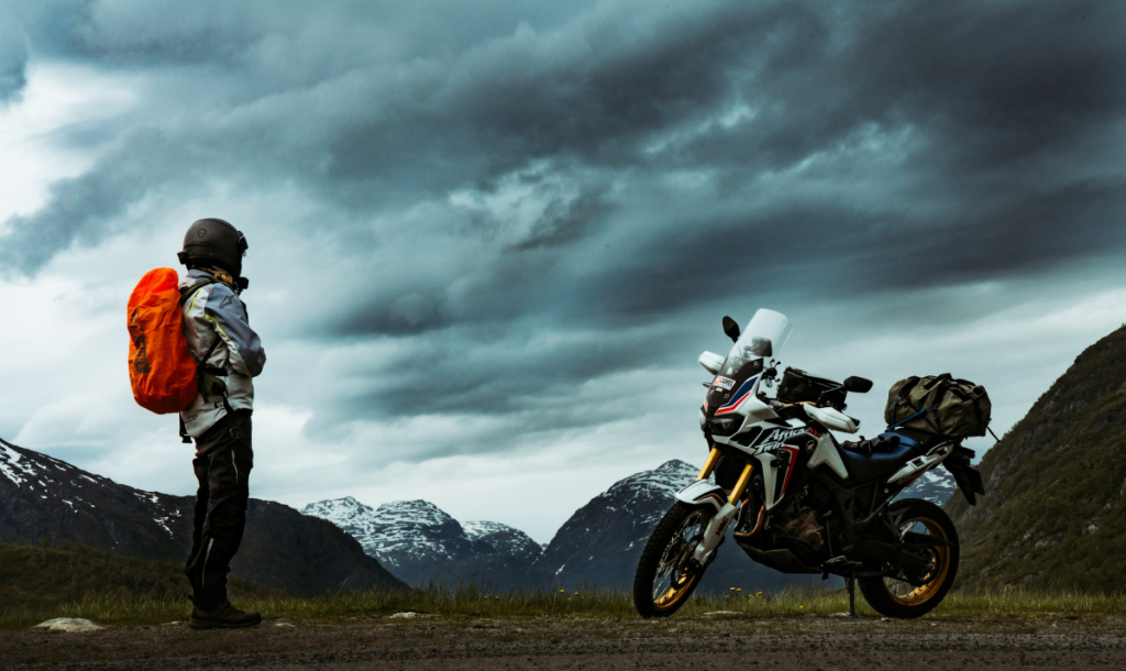 A lone motorcyclist with an orange backpack stands on a mountain road beside a packed adventure bike, gazing at dramatic storm clouds over snow-capped peaks.