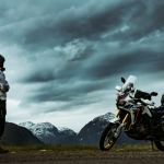 A lone motorcyclist with an orange backpack stands on a mountain road beside a packed adventure bike, gazing at dramatic storm clouds over snow-capped peaks.