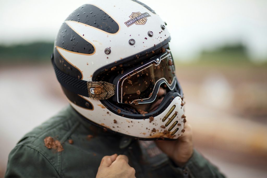 A motorcyclist wearing a Harley-Davidson full-face helmet and goggles is covered in mud and rain, tightening their chin strap.