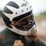 A motorcyclist wearing a Harley-Davidson full-face helmet and goggles is covered in mud and rain, tightening their chin strap.
