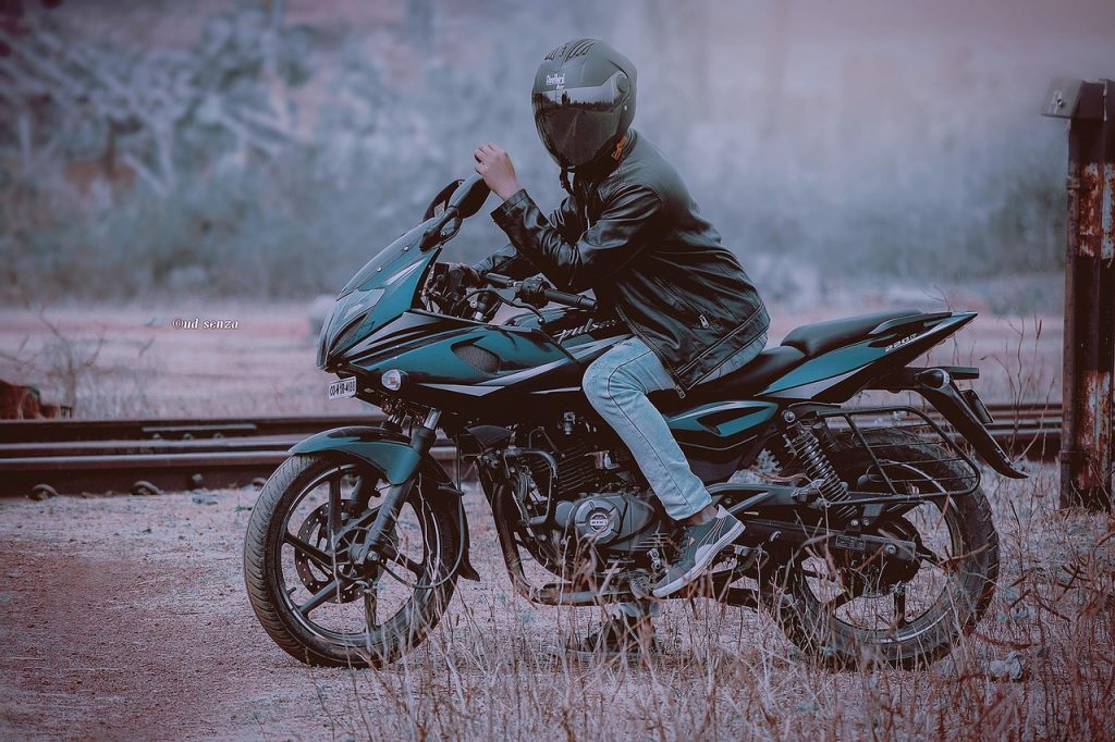 A motorcyclist wearing a black helmet and leather jacket sits on a Bajaj Pulsar near train tracks in a muted outdoor setting.