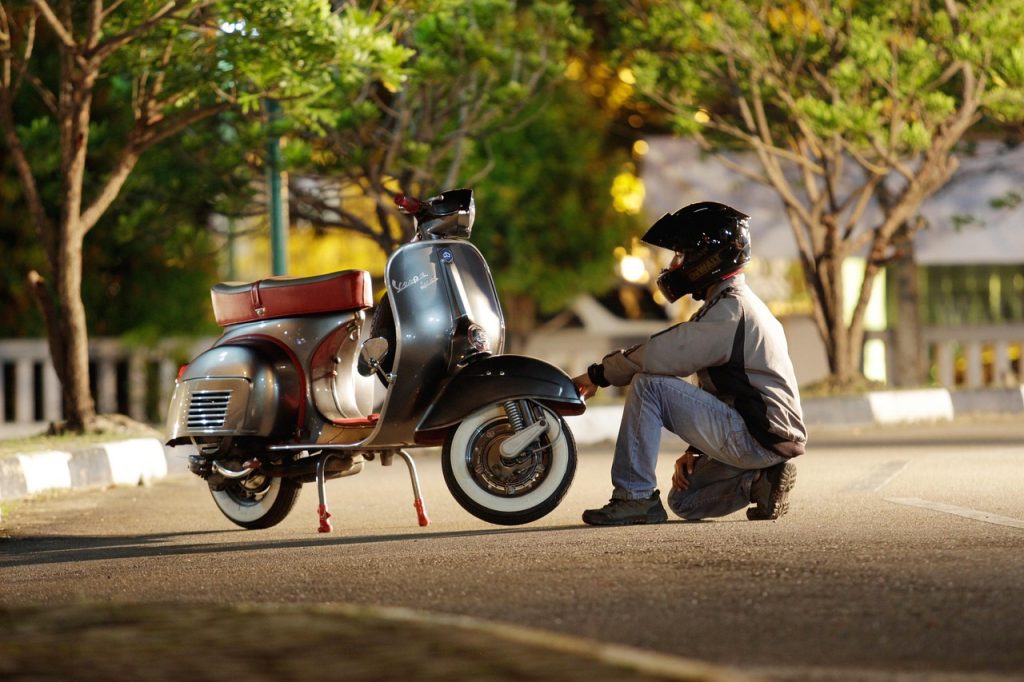 A rider wearing a helmet crouches beside a vintage scooter under warm street lighting, surrounded by trees and a quiet urban setting.