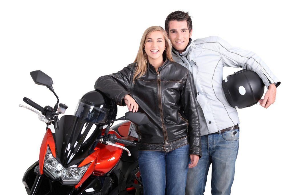 A smiling couple dressed in motorcycle gear stands beside a red and black sport bike, ready for a ride.







