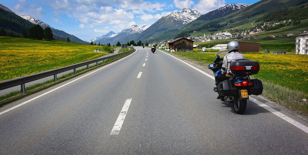 A motorcyclist rides along a scenic, open highway flanked by green fields and snow-capped mountains under a partly cloudy sky, with another rider visible ahead in the distance.
