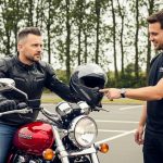 A motorcycle instructor hands a helmet to a rider during a training session in a controlled parking lot environment.