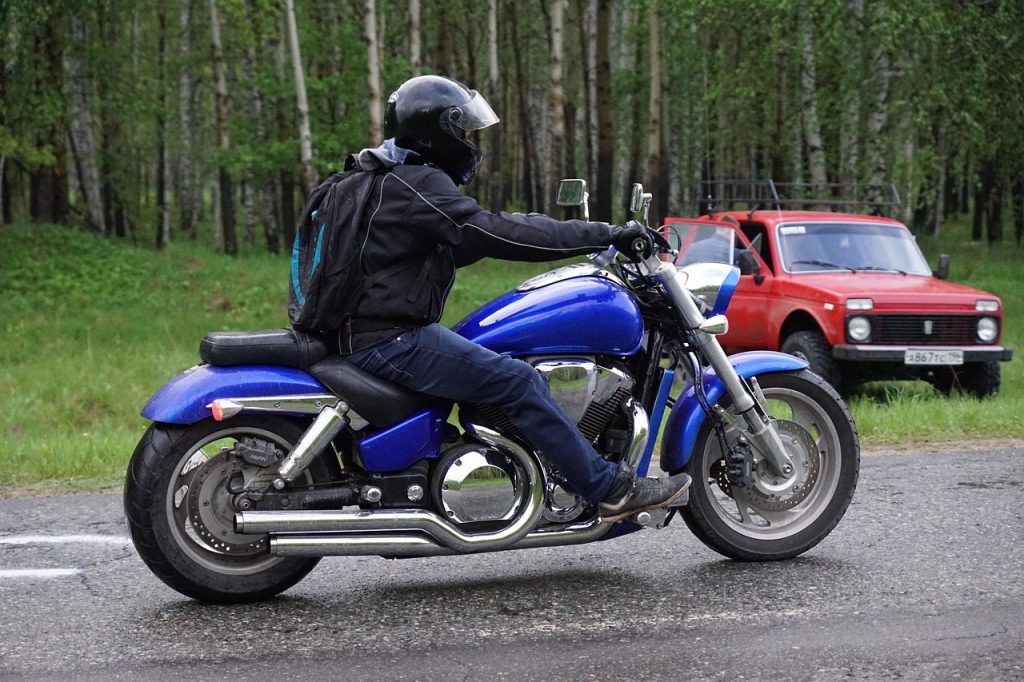 A motorcyclist rides a blue cruiser on a wet road, fully geared in a black waterproof jacket, helmet, gloves, and backpack—demonstrating the importance of wearing rain-ready riding gear to stay comfortable and dry when the rain returns.