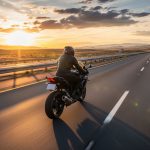 A person on a black motorcycle rides down an empty highway towards the setting sun, with mountains visible in the distance.