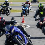 A group of motorcyclists is practicing various riding skills, including cornering, wheelies, and stops, on a closed asphalt course marked with cones.