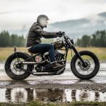 A rider wearing a helmet and leather jacket cruises on a custom-style motorcycle along a wet rural road, with puddles reflecting the bike and a misty landscape in the background.