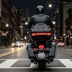 Rear view of a motorcyclist in a black leather jacket riding a touring bike with glowing red tail lights and a New York license plate on a city street at night.
