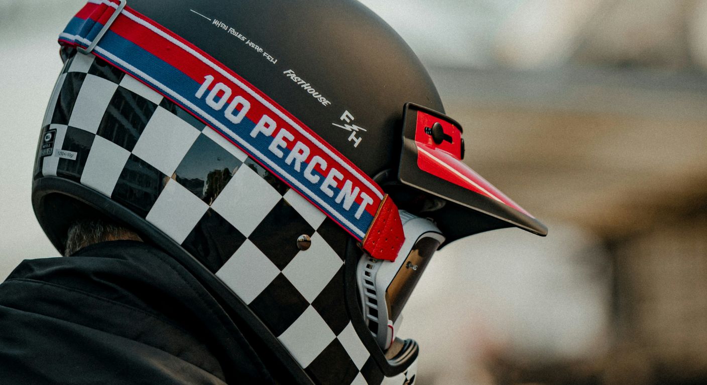 Close-up of a motorcycle rider wearing a black helmet with a checkered pattern and “100 PERCENT” text, viewed from the side.