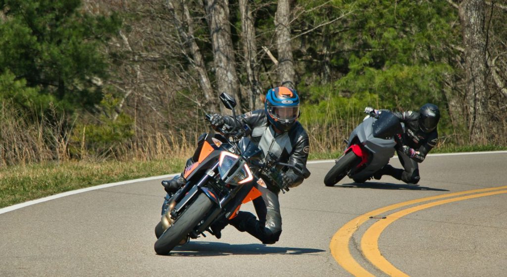 Two motorcyclists in full riding gear leaning aggressively into a curve on a winding road, with the lead rider on an orange street bike and a second rider following closely behind.