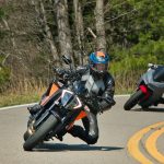 Two motorcyclists in full riding gear leaning aggressively into a curve on a winding road, with the lead rider on an orange street bike and a second rider following closely behind.