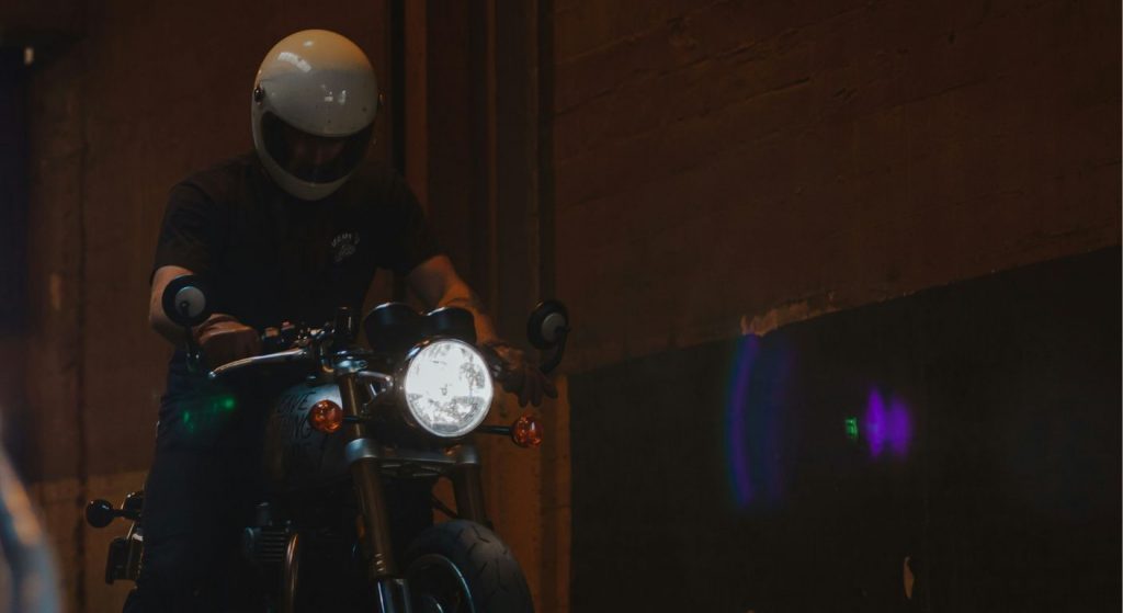Motorcyclist in a white helmet rolling a bike into a dimly lit garage or tunnel, with the headlight illuminating the space.
