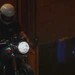 Motorcyclist in a white helmet rolling a bike into a dimly lit garage or tunnel, with the headlight illuminating the space.